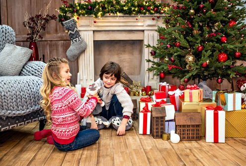 A young girl and boy opening presents under the Christmas tree fireplace with stockings hung
