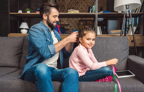 smiling father braiding his smiling daughters hair on the sofa as she plays with ribbons