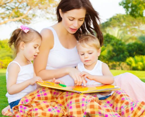 Image of cute young female with two little children read book outdoors, brother and sister with mother learning in the park in spring time, preschool education, day care, happy family concept